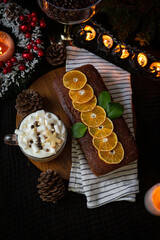 Festive homemade cake topped with sliced oranges, served with hot chocolate with marshmallows, surrounded by Christmas decorations.