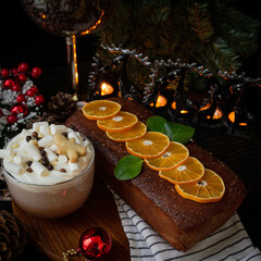 Festive homemade gingerbread cake adorned with orange slices and green leaves, accompanied by a hot cocoa topped with marshmallows and cookies.