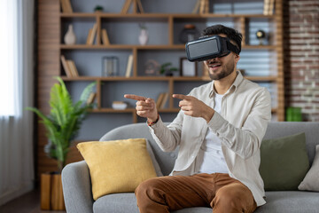 Man enjoying virtual reality experience using VR headset on sofa in cozy living room setting,...