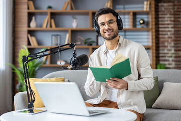 Young man hosting a podcast from home with professional microphone and laptop while reading a book, promoting knowledge sharing and engaging content creation.