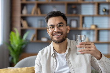Smiling man with glasses holding glass of water in cozy indoor setting, radiating positivity and health