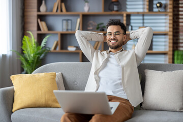 Relaxed man enjoying leisure time on couch with laptop in cozy living room, feeling content and stress-free