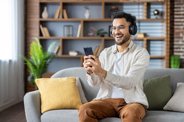 Smiling man enjoying music on headphones while using smartphone at home, relaxing on sofa. Tech-savvy individual immersed in digital entertainment.
