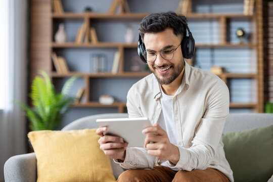 Smiling young man with headphones using digital tablet on couch in modern living room for relaxation
