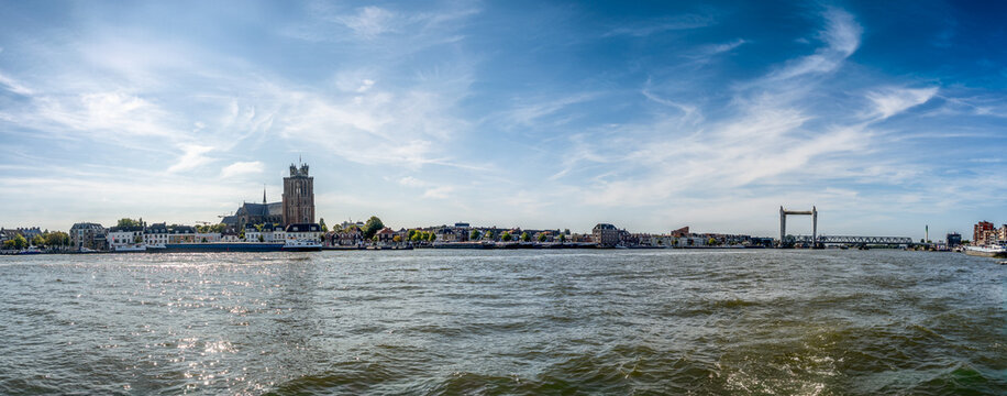 A panoramic view of the riverside city Dordrecht featuring a prominent church and a modern bridge against a clear blue sky. Small buildings and greenery line the water's edge, adding charm to the view