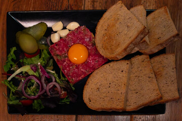 Rustic steak tartare with raw egg yolk, sliced bread, pickles, garlic and fresh salad on a wooden table.