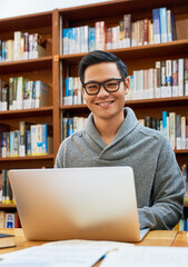 Laptop, portrait and man in library for learning with university, education and scholarship with smile. Books, online research and happy student on campus studying for exam, knowledge and growth