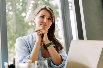 Smiling, attractive woman freelancer working on laptop in cafe looking at camera