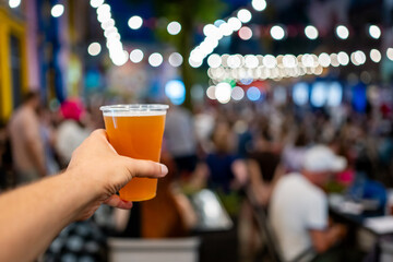 Hand holding a plastic cup of beer with a blurred festival background. Bokeh lights create a lively atmosphere, capturing the essence of a fun outdoor event. Perfect for themes of celebration