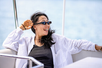 A woman of Asian ethnicity in a white shirt relaxes on a boat on a sunny day, smiling warmly. The...