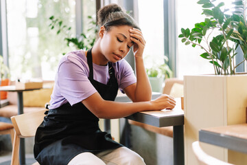 Young African American tired waitress holding her head while sitting in coffee shop