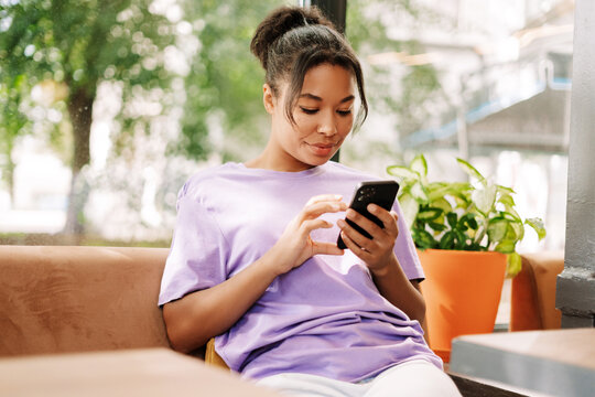 Young African American woman using smartphone in a cafe, browsing social media - Powered by Adobe
