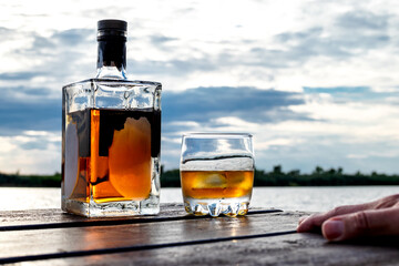A chilled bottle and a glass of spirits on rocks are on a wooden dock by the water. Heavy clouds at...