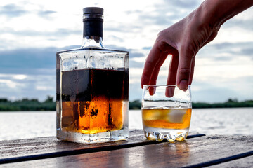 A chilled bottle and a glass of spirits on rocks are on a wooden dock by the water. Heavy clouds at...