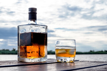 A chilled bottle and a glass of spirits on the rocks are on a wooden dock by the water. Heavy clouds at sunset in the background.