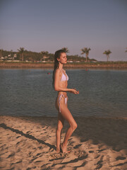 Woman in white bikini standing on beach next to body of water on sunny day with clear blue sky