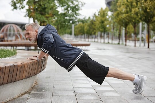 Side view of fit elderly man in sportswear doing push ups on bench working out regularly in city park with tiles, copy space - Powered by Adobe
