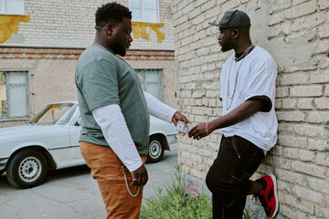 African American man giving wad of cash to guy who leaning on old garage