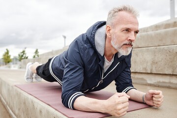 Sporty determined elderly man holding plank position on yoga mat strengthening core muscles during...