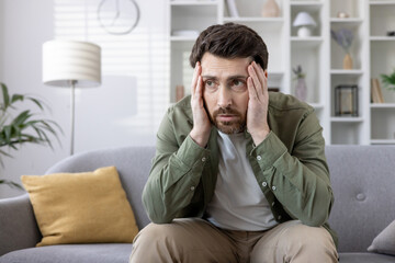 Concerned man seated on sofa holding head, depicting stress and anxiety in domestic setting....