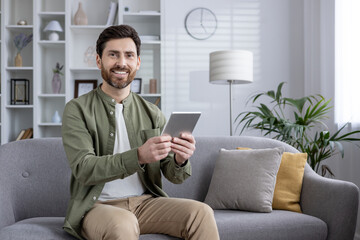 Confident man wearing green shirt sits comfortably on sofa using tablet. Living room setting conveys modern lifestyle and relaxation. No background description included in keywords.