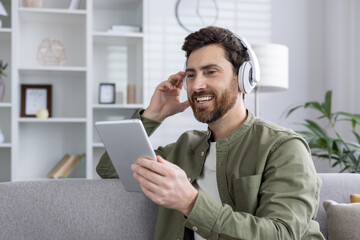 Man with headphones using tablet while relaxing on couch. Smiling, enjoying music or podcast in bright living room. Casual home environment promotes comfort and leisure.