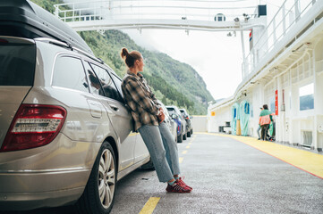 Woman leans against her car on Norwegian ferry surrounded by other vehicles with misty green...