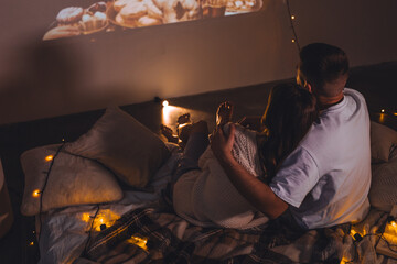 Obraz premium Romantic surprise for girlfriend or boyfriend. Bedroom prepared for watching old movies with popcorn, decorated with lights and candles. Cozy home Christmas atmosphere, hot chocolate with marshmallow