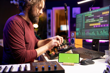 Audio engineer uses knobs on stereo gear for mixing and mastering session, having a smartphone with greenscreen display in home studio. Musician learning to edit tunes with daw software.