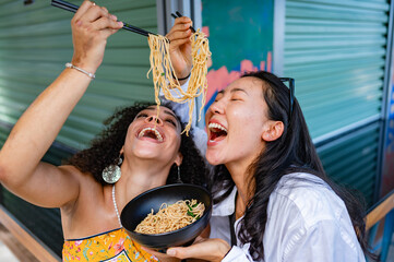 Cheerful girl friends- of Afro-Latin, and  Asian ethnicity, enjoying eating noodles outdoors...