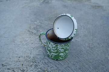 An enamel mug with green and white marbling sits on a gray surface, its lid slightly ajar.