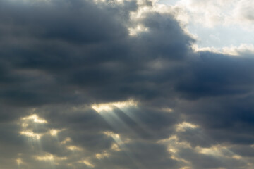 Long rays of sun break through many dark clouds in the blue sky. Cloudy weather. Blue, yellow and white colors. Religion, God, Supreme. Travel, calm, peace.