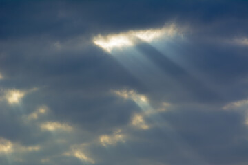 Long rays of sun break through many dark clouds in the blue sky. Cloudy weather. Blue, yellow and white colors. Religion, God, Supreme. Travel, calm, peace.