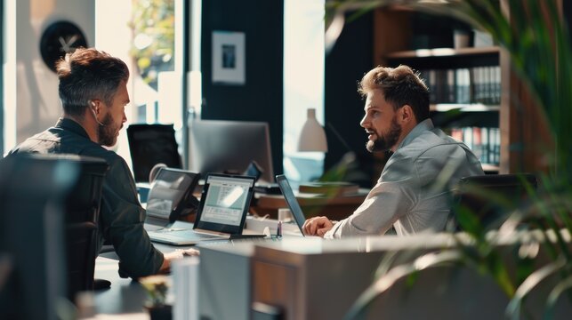 Two businessmen having a discussion while working on laptops in busy open plan office