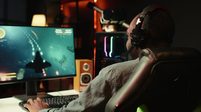 Man playing SF shooter videogame on computer monitor, talking into headset with friends. Gamer using headphones while enjoying game with spaceship flying to chat with Esports team, camera B