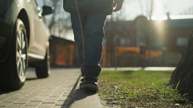A close-up shot of a child walking along a pavement holding a stick, missed his step before climbing back onto the pavement the child is dressed in a shiny black jacket, surrounded by