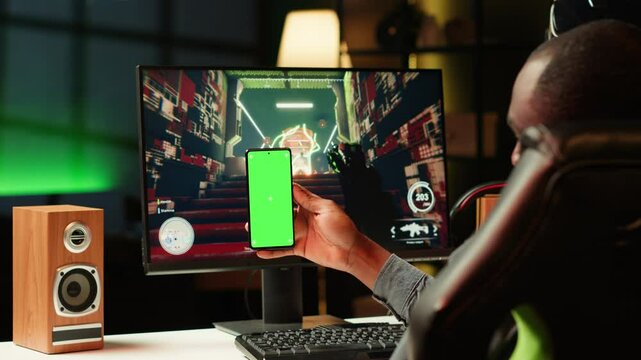 Man texting friends on green screen phone while playing first person shooter videogame at home. Gamer looking at mockup smartphone display, enjoying online discussion with mates