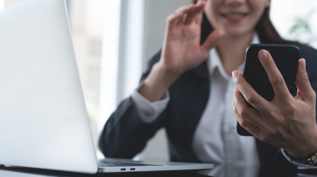 Young asian businesswoman smiling and using mobile phone for video calling with colleague, online meeting via mobile app during working on laptop computer at modern office, webinar