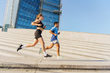 A fit couple running up urban stairs, with modern architecture towering behind, under a clear blue sky.