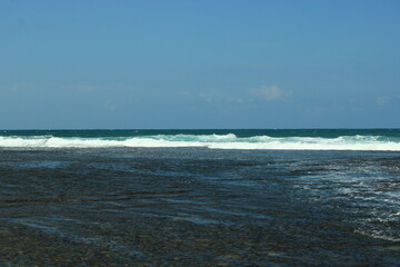 Blue sky over a calm ocean with waves crashing on a rocky shore.
