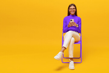 Woman spending leisure time surfing mobile internet sitting on chair on yellow copy space background