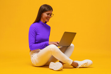 Side view portrait of woman browsing social media on laptop sitting on floor on yellow background