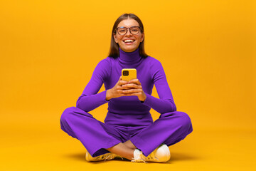 Laughing woman sitting on floor on yellow background browsing social media posts on phone