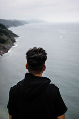 Fototapeta premium chico joven de pelo rizado negro mirando el mar desde un acantilado en un día de tormenta