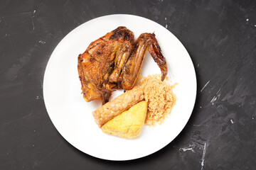 A plate of savory dishes including sweet grilled chicken with a dark brown color, fried tofu and tempeh, and crispy crumbs, served on a white plate against a dark background.