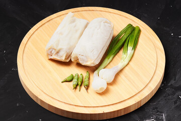 Two frozen lumpia filled with bamboo shoots and meat, with white wrappers, ready to fry. Served with green chili peppers and scallions, photographed on a round wooden tray against a dark background.