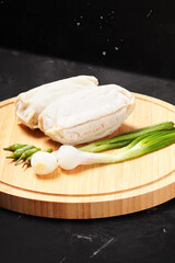 Two frozen lumpia filled with bamboo shoots and meat, with white wrappers, ready to fry. Served with green chili peppers and scallions, photographed on a round wooden tray against a dark background.