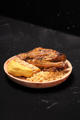A plate of savory dishes including sweet grilled chicken with a dark brown color, fried tofu and tempeh, and crispy crumbs, served on a wooden plate against a dark background.