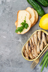 Tin can with sprats, bread, vegetables and herbs on a dark background, top view.