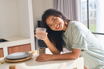 Smiling Woman Relaxing In Bright Kitchen, Holding Glass Of Water. Morning Routine, Hydration,...
